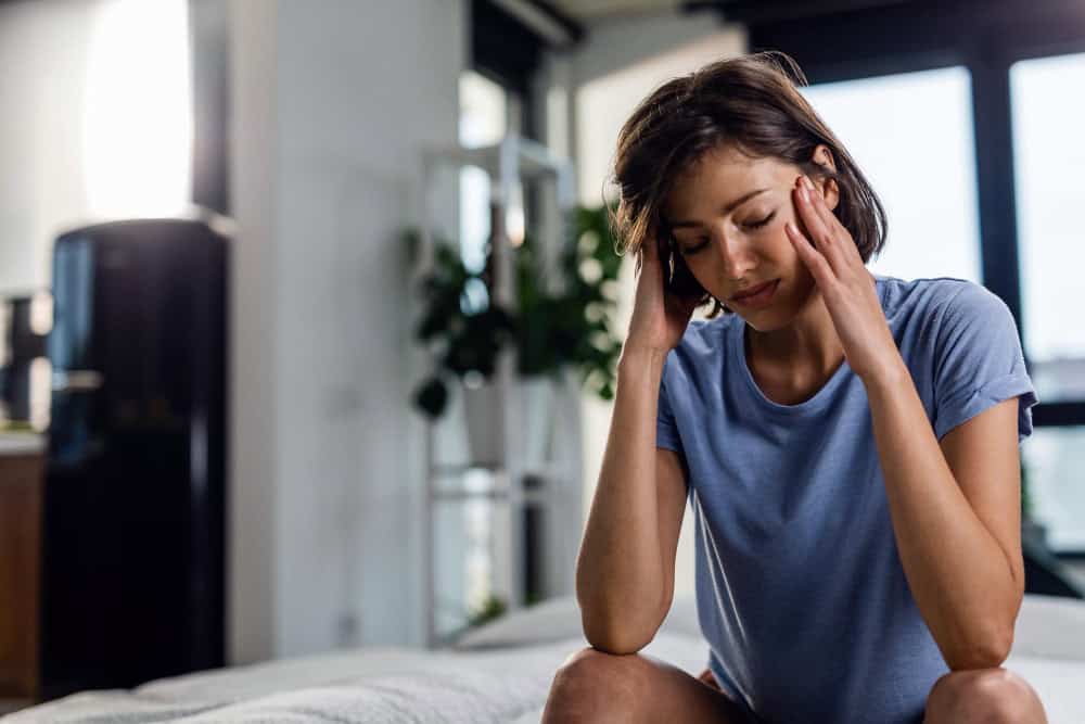 Sad woman suffering from headache and sitting on the bed with eyes closed while holding her head in pain 